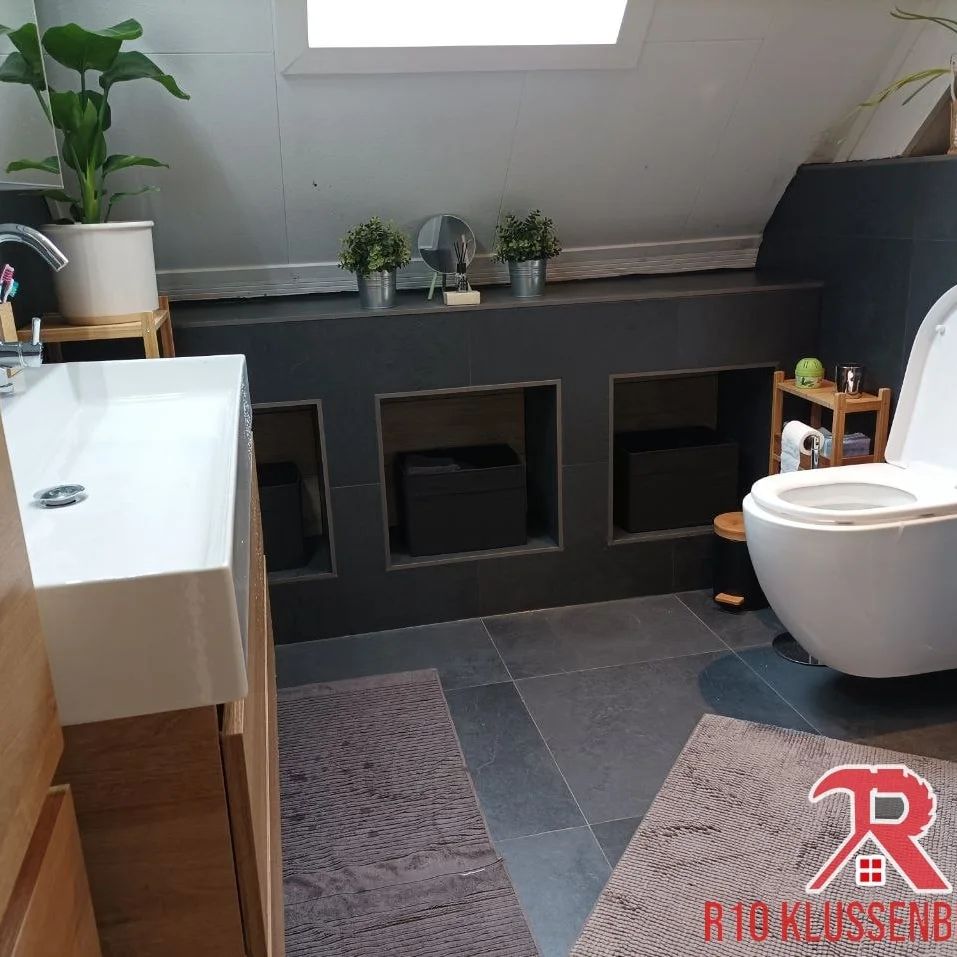 Zolderbadkamer met donkere tegels, nissen in de wand, een hangend toilet en planten