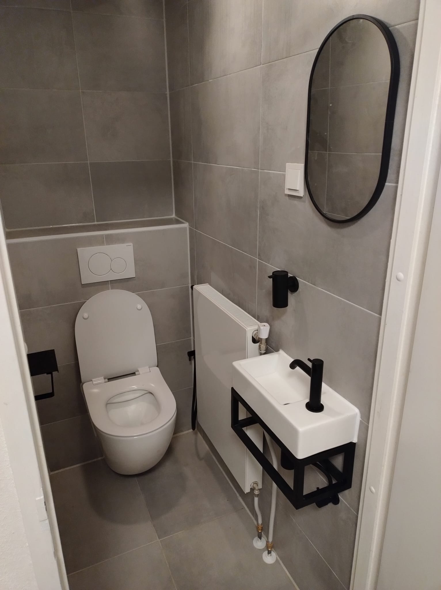 Verbouwd toilet met grijze tegels, een zwarte ovale spiegel en een zwart fonteintje