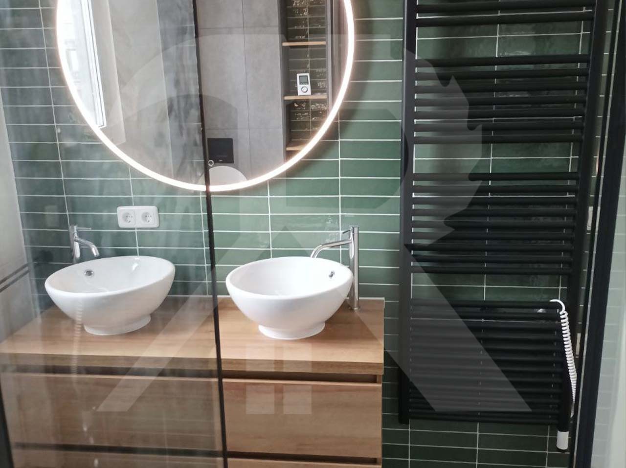 Badkamer met groene tegels, een ronde verlichte spiegel en twee waskommen op een eiken meubel